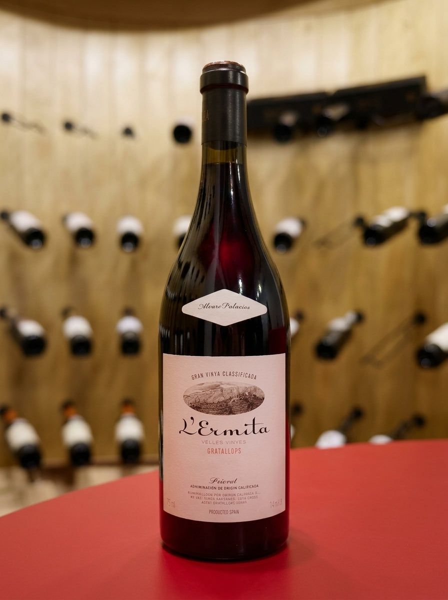 L'ERMITA, PRIORAT red wine bottle displayed on a red table in a wood paneled wine cellar with bottles lining the shelves.
